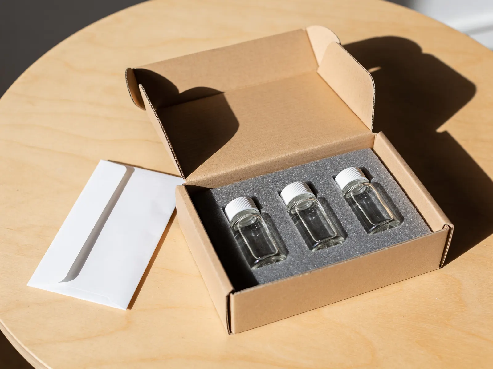 An open kraft shipping box holding three capped glass vials in grey foam, beside a white envelope